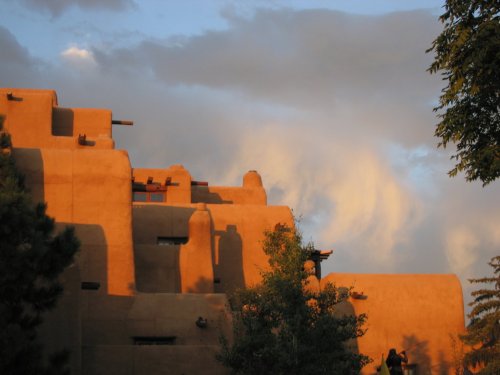 Adobe-style buildings bathed in warm sunset light, with clouds and trees in the background.