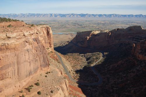 A panoramic view of a canyon with a winding road, surrounded by rocky cliffs and distant mountains under a clear sky.