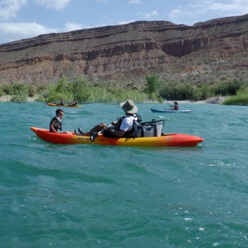 A person in a kayak on a river, surrounded by others kayaking, with rocky cliffs and greenery in the background.