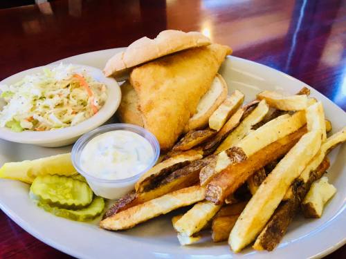 A plate with a fried fish sandwich, fries, coleslaw, pickles, and a side of tartar sauce.