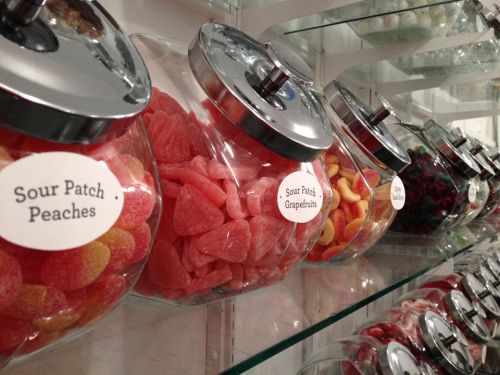 A display of glass jars filled with colorful gummy candies, labeled with flavors like Sour Patch Peaches and Grapefruits.