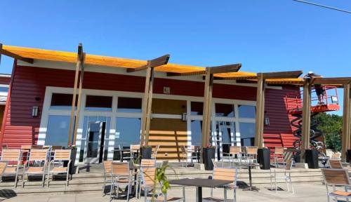 A modern restaurant exterior with a wooden awning, outdoor seating, and a worker on a lift. Clear blue sky above.