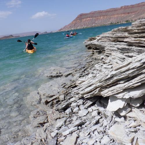 A person kayaking near a rocky shoreline with layered stones, surrounded by blue water and a mountainous landscape.