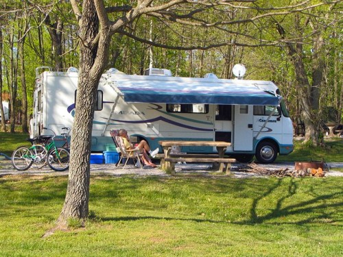 A parked RV in a green campsite, with a picnic table, a bicycle, and a person relaxing in a chair nearby.
