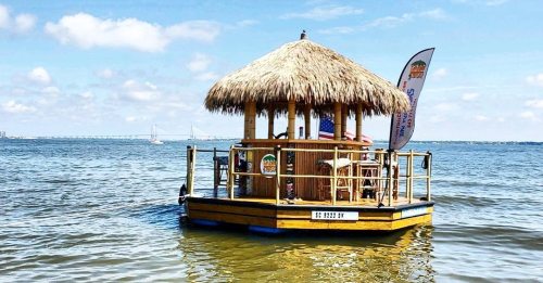 A small tiki hut on a floating platform in calm water, with flags and a scenic sky in the background.