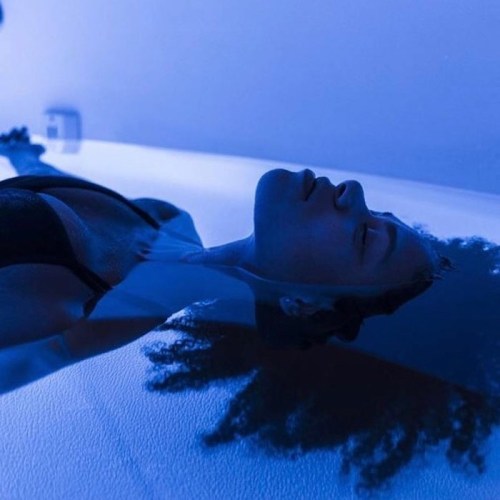 A person floats in a blue-lit space, eyes closed, with their hair spread out in the water.