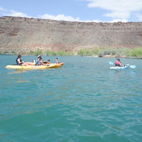 Two kayakers paddle on a turquoise lake, surrounded by rocky cliffs and greenery under a bright blue sky.