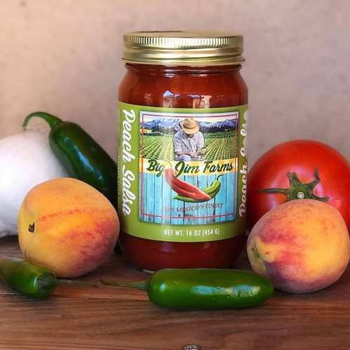 A jar of Big Jim Farms peach salsa surrounded by fresh peaches, jalapeños, and a tomato on a wooden surface.