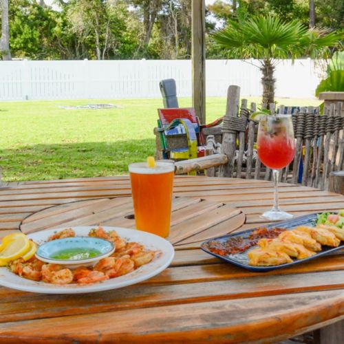 A wooden table outdoors with shrimp, a cocktail, and a beer, surrounded by greenery and a white fence.