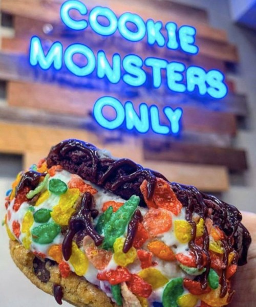 A colorful cookie sandwich filled with ice cream and topped with chocolate drizzle, in front of a "Cookie Monsters Only" sign.