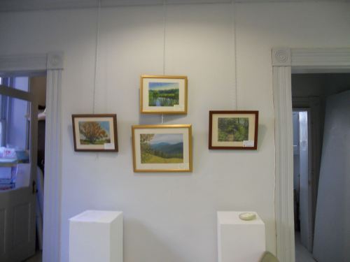 Four framed landscape paintings hang on a white wall, with two pedestals visible in the foreground.