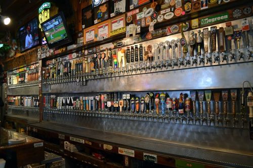 A bar with a long row of beer taps, various brands and labels displayed, and a TV showing a sports game in the background.