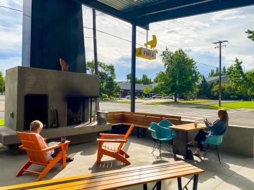 A cozy outdoor seating area with a fireplace, two people relaxing, and a sign for "Finch" in a sunny setting.