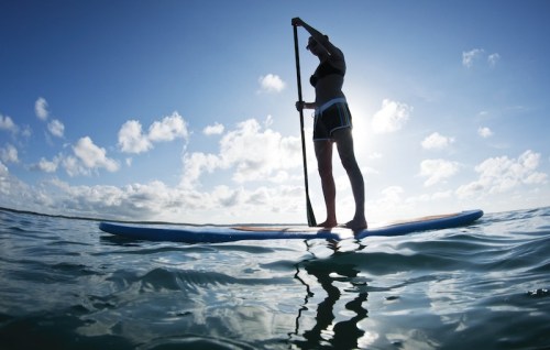 Silhouette of a person paddleboarding on calm water under a bright blue sky with fluffy clouds.