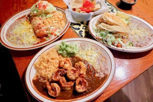 Three colorful plates of Mexican food featuring shrimp, enchiladas, tacos, rice, beans, and guacamole.