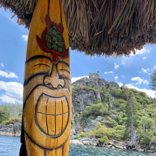 A carved wooden tiki mask in the foreground with a scenic view of rocky hills and trees in the background.