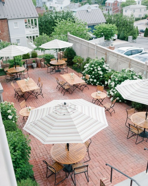A charming outdoor patio with wooden tables, chairs, and large umbrellas surrounded by greenery and blooming flowers.