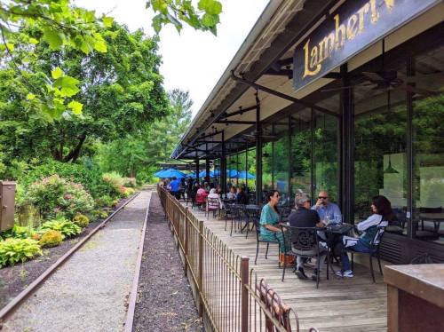 Outdoor dining area of a restaurant with patrons seated at tables, surrounded by greenery and blue umbrellas.