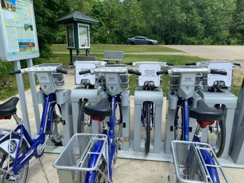 A bike-sharing station with several blue bicycles parked and a map kiosk in a green outdoor area.