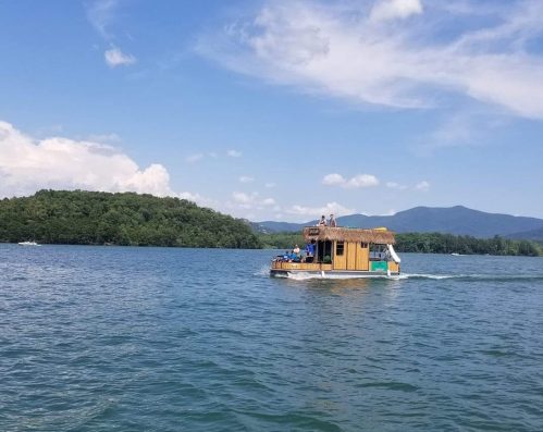 A small houseboat glides across a calm lake under a blue sky with fluffy clouds and green hills in the background.
