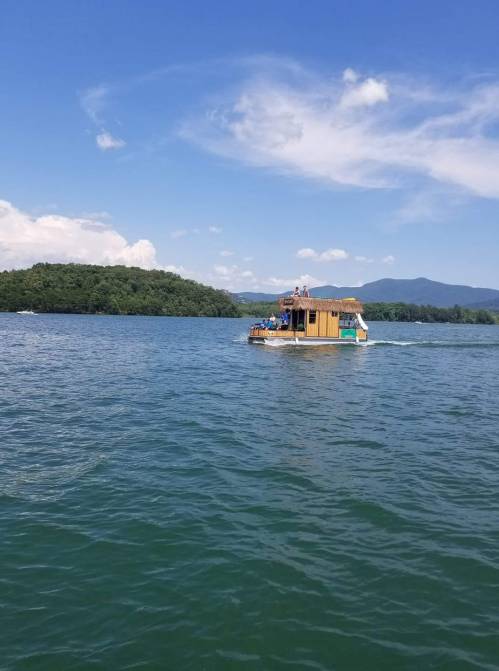 A houseboat glides across a calm lake under a blue sky with fluffy clouds and green hills in the background.