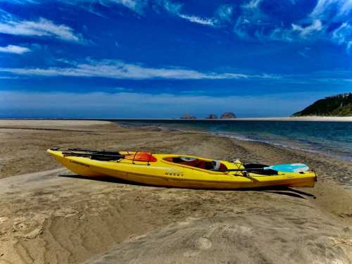A yellow kayak rests on a sandy beach with blue water and a clear sky in the background.