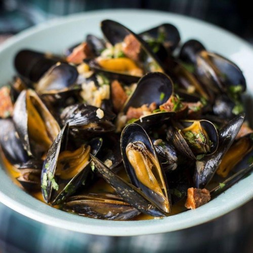 A bowl of mussels cooked with herbs and spices, garnished with bits of meat and served in a light broth.