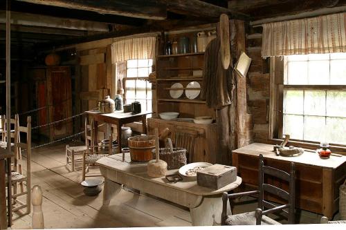 A rustic interior of a wooden cabin, featuring a table, chairs, shelves with dishes, and various vintage kitchen items.