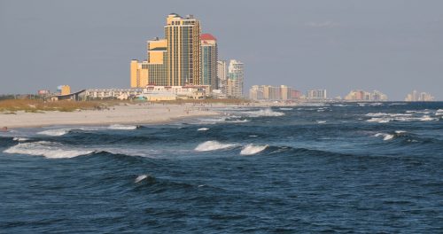 A coastal view featuring waves, sandy beach, and tall buildings along the shoreline under a clear sky.