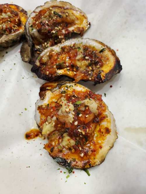 Grilled oysters topped with a spicy, savory mixture and garnished with herbs and spices on a white surface.