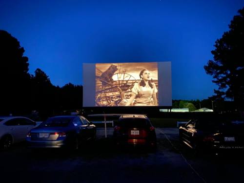 A drive-in movie screen shows a classic film scene at dusk, with parked cars in the foreground.