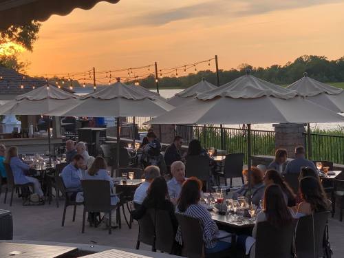 Outdoor dining scene at sunset, with people seated under umbrellas by a lake, enjoying a relaxed atmosphere.
