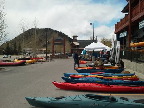 Colorful kayaks lined up along a street near a mountain, with tents and people preparing for water activities.