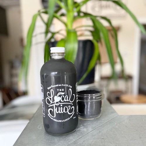 A dark juice bottle labeled "The Local Juice" next to a small jar, with a green plant in the background.