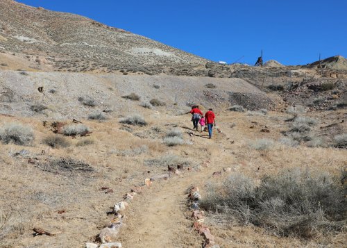 Two hikers walk up a dirt path through a dry, rocky landscape under a clear blue sky.