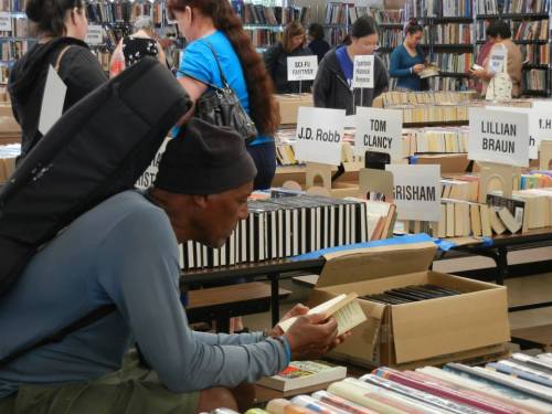 A man examines a book at a busy book sale, surrounded by tables filled with various titles and people browsing.