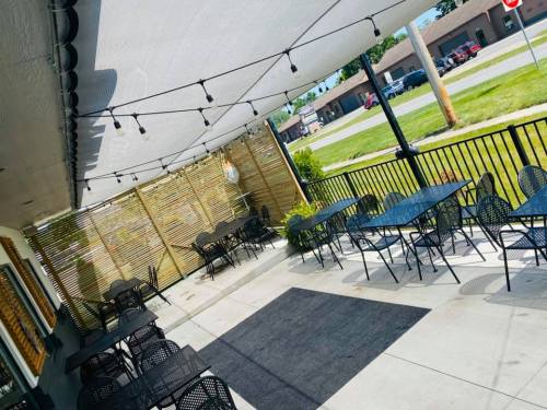 Outdoor patio with black metal tables and chairs, shaded by a canopy and string lights, surrounded by greenery.
