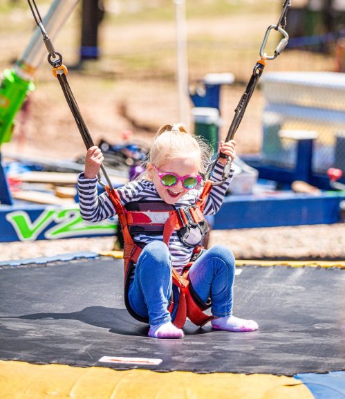 A young girl in sunglasses enjoys bouncing on a trampoline, secured in a harness with safety straps.