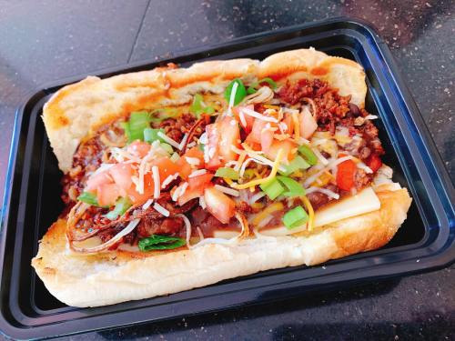 A loaded chili cheese dog topped with chopped tomatoes, green onions, and shredded cheese in a toasted bun.