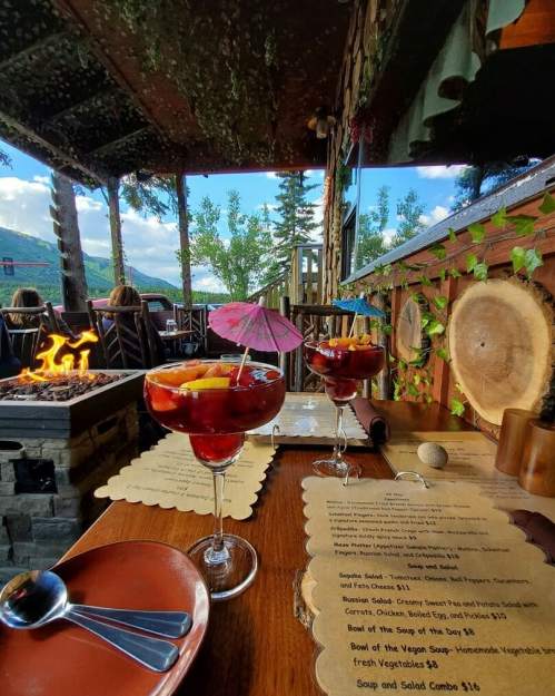 Two colorful cocktails with fruit and umbrellas sit on a wooden table, with a scenic view and a menu in the background.