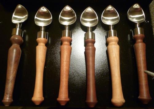 A row of six ice cream scoops with wooden handles in various shades, set against a dark background.