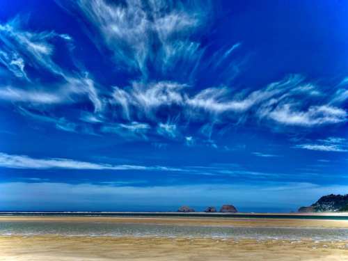 A serene beach scene with a vast blue sky, wispy clouds, and distant rock formations on the horizon.