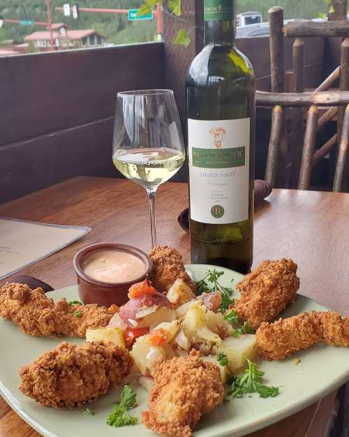 A plate of fried chicken with salad, a glass of white wine, and a bottle of Chardonnay on a wooden table.