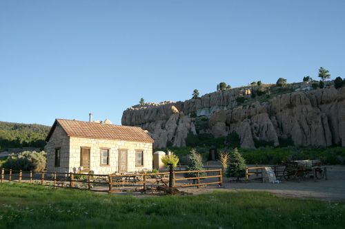 A rustic stone house near rocky cliffs, surrounded by greenery and a wooden fence under a clear blue sky.