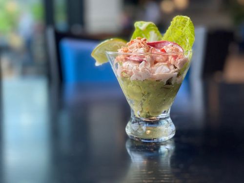 A glass dish filled with shrimp, avocado, and lettuce, garnished with lime and radish, on a dark table.