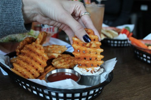 A hand holding a waffle fry above a basket of fries and dipping sauce on a wooden table.
