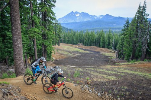 Two mountain bikers navigate a dirt trail through a forest, with mountains visible in the background.