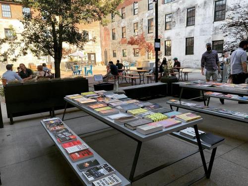 Outdoor scene with tables displaying books, people socializing in the background, and a rustic building.
