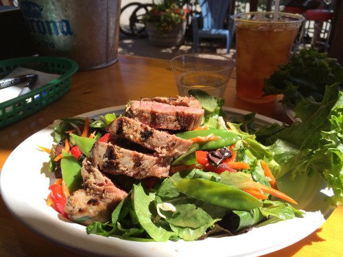 A plate of sliced grilled steak on a bed of mixed greens, topped with red peppers and snap peas, with a drink in the background.