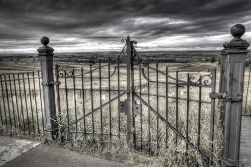 A wrought iron gate stands open, leading to a vast, grassy landscape under a moody, overcast sky.